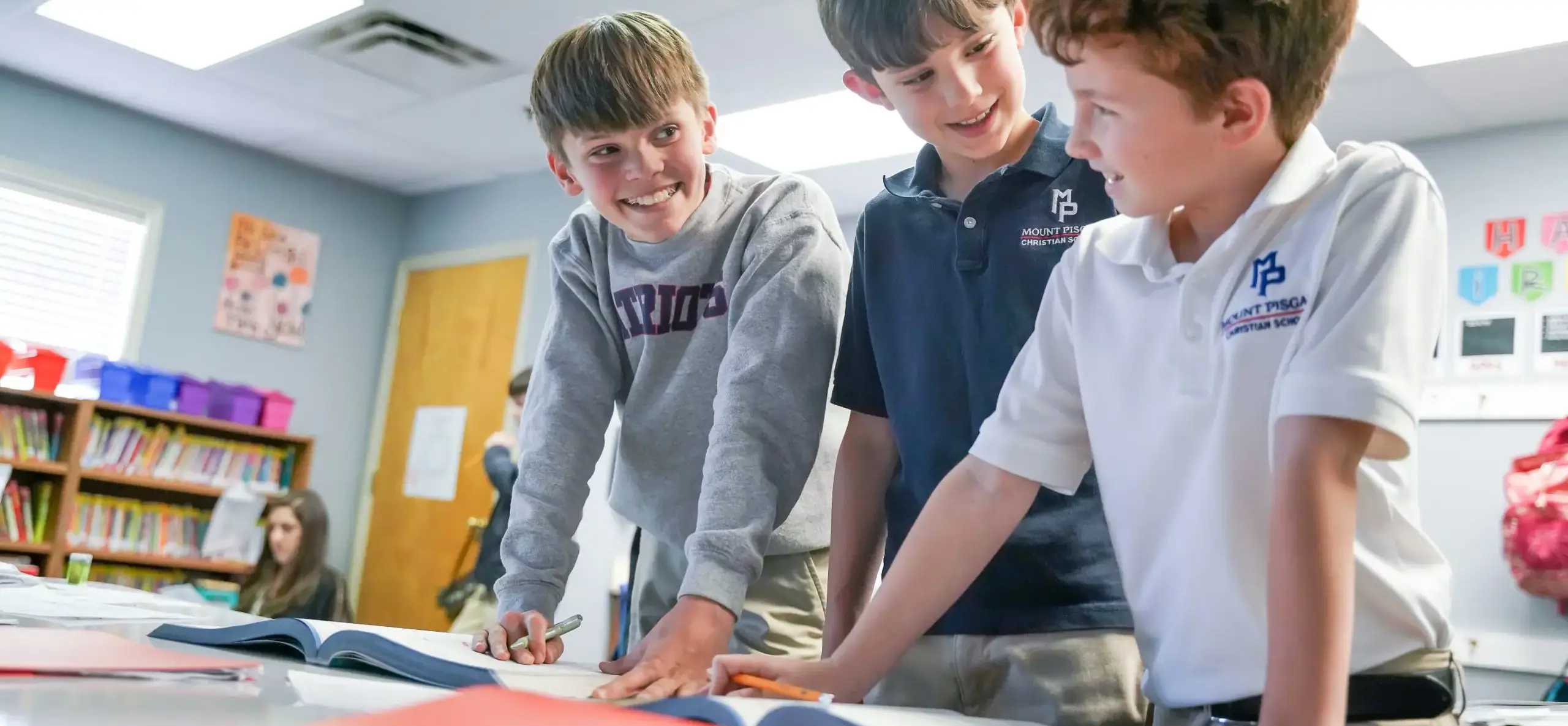 Three students stand near a desk, working in a classroom